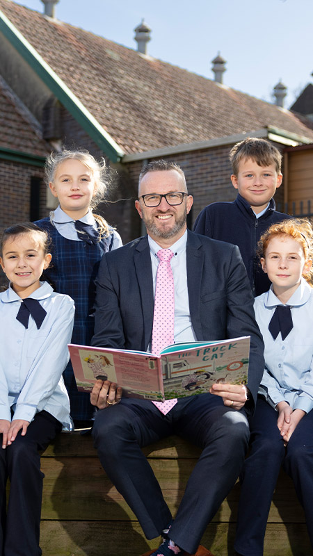 Matthew Bond, Principal of Our Lady of the Nativity Primary Lawson, reading to students in an outdoor setting.