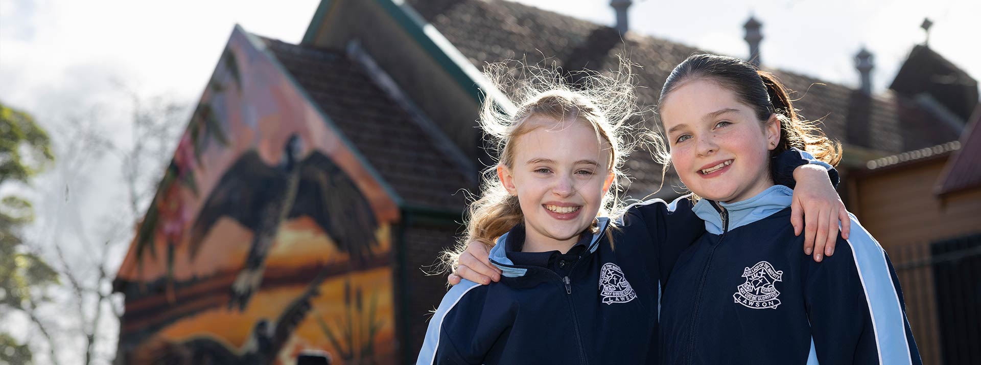 Two smiling OLN Lawson students outdoors. The Church and Mural is in the background.