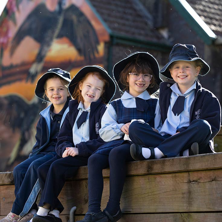 Four Stage 1 (Kindy to Year 1) Our Lady of the Nativity Lawson students sitting outdoors. The Church and Mural is in the background.